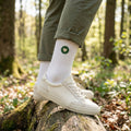 Weisse Premium Socken aus Bio-Baumwolle mit aufgesticktem grünen Herzlogo. Im Hintergrund ist Wald zu sehen.