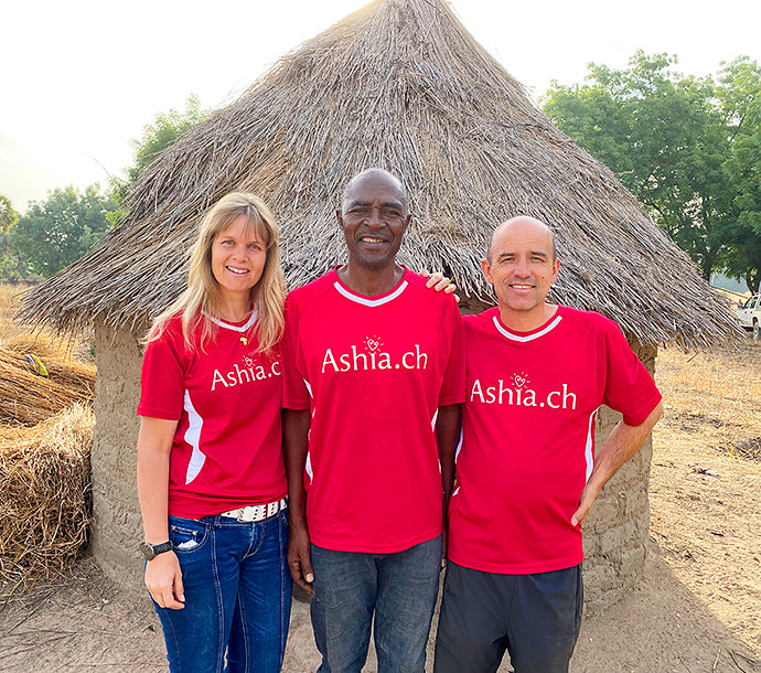 Katja und Felix Bruhin von Ashia Kamerun zusammen mit Gregory vor einem kamerunischen Lehmhaus.
